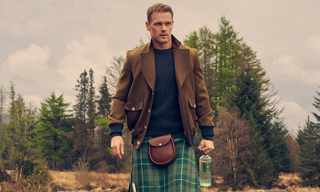 Sam Heughan in a brown jacket and green kilt standing in a forested area holding a bottle of The Sassenach Wild Scottish Gin.